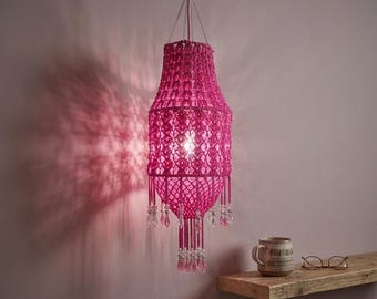 May include: A bright fuchsia macramé pendant light fitting featuring an intricate knotted cord pattern, suspended by silver chains. The lower section is adorned with dangling clear crystal-like beads, casting a patterned shadow on the wall.