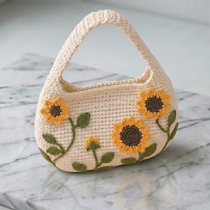 May include: A cream-coloured crochet handbag featuring a single handle and a decorative pattern of three yellow sunflowers with dark brown centres and green leaves stitched onto the front of the bag. The bag has a rounded, soft-sided structure.