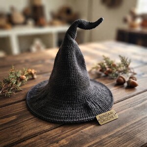 May include: A dark grey crocheted pointed hat with a wide brim and a curved top. A small rectangular metal tag attached to the brim features the text SUNDARI CROCHET. The hat is constructed from a dense, textured yarn stitch pattern.