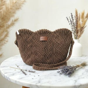 May include: A dark brown crochet shoulder bag featuring a textured leaf-like pattern throughout the exterior. The bag includes a matching crochet strap and a small rectangular brown leather-like label centered on the front with text.