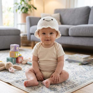 Può includere: Un cappello a secchiello all'uncinetto bianco caratterizzato da un semplice viso ricamato con due occhi neri e una bocca curva. Il cappello ha una struttura in filato morbido e testurizzato con una tesa leggermente ondulata.