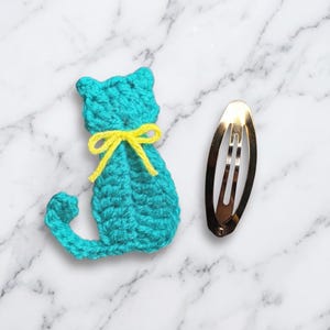 May include: A turquoise crocheted cat applique featuring a bright yellow yarn bow tied around its neck, positioned next to a shiny, oval-shaped silver metal hair clip. The cat figure has a curved tail and pointed ears, crafted from yarn.
