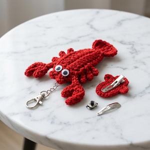 Peut inclure: Un porte-clés homard en crochet rouge avec des yeux mobiles attaché à un mousqueton en métal argenté. À côté se trouve une petite pièce en crochet rouge avec une pince à cheveux en métal argenté, et une pince à cheveux métallique séparée.
