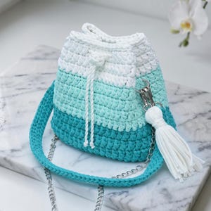 Pode incluir: Uma bolsa de crochê tipo saco com um gradiente de cores branco, menta claro e azul-petróleo escuro. A bolsa inclui um fecho de cordão branco, uma borla branca a condizer presa a um clipe de metal prateado e uma alça de crochê azul-petróleo.