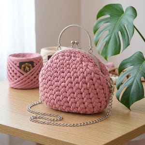 Könnte beinhalten: Eine altrosa gehäkelte Handtasche mit dicker, geflochtener Garnstruktur, einem silberfarbenen Metall-Kussverschluss, einem passenden silberfarbenen Metallgriff und einem abnehmbaren silberfarbenen Metallkettenschulterriemen.