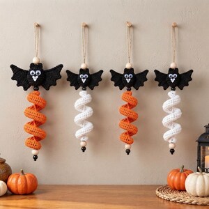 May include: Four hanging crochet bat ornaments, each featuring a black bat head with googly eyes and outstretched wings. Below each bat is a long, spiraling crochet tail in either bright orange or white, accented with small wooden and black beads.
