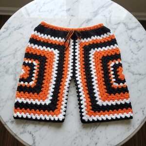 May include: Crochet shorts featuring a granny square pattern in alternating bands of black, white, and orange yarn. The shorts have an elasticized waistband with a thin, matching black and orange braided drawstring tie at the center front.