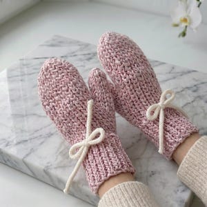 Può includere: Un paio di muffole lavorate a maglia color rosa chiaro con un motivo a maglia testurizzato e un polsino a coste. Ogni muffola è decorata con un piccolo cordoncino color bianco sporco annodato a fiocco sul polso.