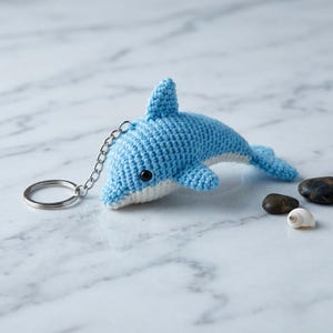 May include: A light blue and white crocheted dolphin keychain featuring a small metal ring attached by a short silver chain. The dolphin has a smooth, textured yarn surface, a single visible black eye, a dorsal fin, and a tail fin.