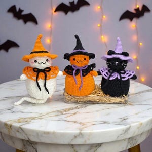 May include: Three crocheted figures featuring a white ghost with an orange hat, an orange pumpkin with a black hat, and a black cat with a lavender hat. Each figure wears a decorative collar with a bow and has embroidered facial features.