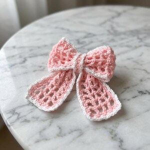 May include: A handmade crochet bow featuring a light pink open-weave grid pattern with a solid white border trim. The bow has a central knot and two long, dangling tails, showcasing a delicate, airy texture throughout the entire piece.