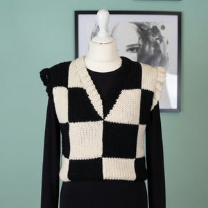 May include: A sleeveless knit sweater vest featuring a bold black and cream checkerboard pattern. The garment has a V-neckline with a thick ribbed border and ruffled shoulder details, displayed on a mannequin over a solid black long-sleeve shirt.