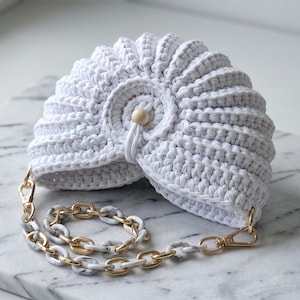 Könnte beinhalten: Eine weiße gehäkelte Handtasche mit muschelartiger, plissierter Textur und einem zentralen Holzperlenverschluss. Die Tasche hat einen abnehmbaren Kettenriemen aus abwechselnden goldfarbenen Metallgliedern und weiß marmorierten Acrylgliedern.