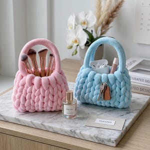 Puede incluir: Dos pequeñas cestas de almacenamiento de punto grueso con asas, una en rosa pastel y otra en azul claro. La cesta rosa contiene brochas de maquillaje, mientras que la azul contiene auriculares inalámbricos blancos, un tubo pequeño y llaves.