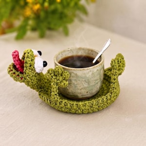 Puede incluir: Un posavasos de ganchillo verde con forma de rana, boca abierta, lengua rosa y grandes ojos blancos con pupilas negras. El posavasos sostiene una taza de cerámica moteada con líquido oscuro y una pequeña cuchara de plata en su interior.