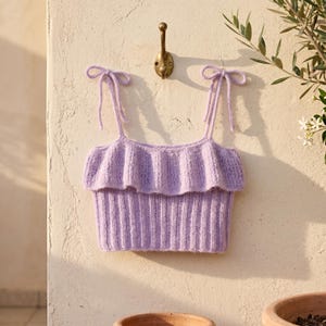 May include: A light purple knit crop top featuring a ribbed texture, a ruffled overlay across the chest, and two thin shoulder straps that tie into bows at the top. The garment is constructed with a soft yarn material and has a straight neckline.