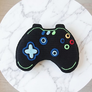 May include: A black crocheted cushion shaped like a video game controller. It features a light blue directional pad, two blue circular buttons, and four smaller buttons in yellow, red, green, and blue. Bright green yarn outlines the controller edges.