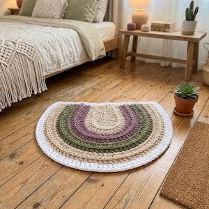 May include: A semi-circular crochet rug featuring concentric bands of color. The design includes a central beige arch, followed by a dusty purple band, an olive green band, a light beige band, and a thick white border along the outer edge.