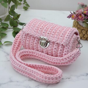 May include: A light pink crochet crossbody bag featuring a textured ribbed pattern, a silver-toned metal push-lock closure, and a matching thick crocheted shoulder strap. The bag has a rectangular shape with rounded edges and a flap closure.