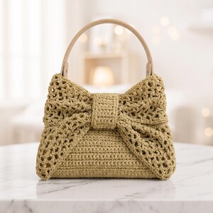 May include: A beige crochet handbag shaped like a large bow. The bag features an open-weave texture, a solid center knot, and a smooth, rounded light-colored handle attached to the top. The structure is firm and maintains a distinct bow silhouette.