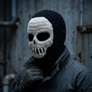 May include: A black crochet balaclava featuring an off-white skull design on the front. The skull pattern includes large dark eye sockets and a detailed jawline with vertical teeth, covering the face while the rest of the head covering is black.