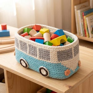 Puede incluir: Una cesta de almacenamiento ovalada de ganchillo diseñada como una furgoneta azul claro y blanca con ventanas grises y ruedas marrón oscuro. La cesta está llena de varios bloques de construcción de madera coloridos, cubos y cilindros.