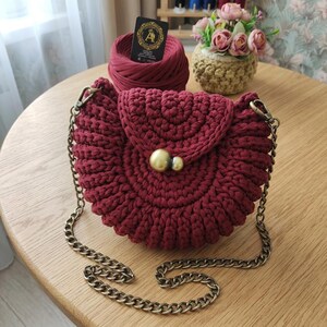 Può includere: Una borsa a mezzaluna lavorata all'uncinetto in rosso scuro, caratterizzata da una struttura in filato spesso e testurizzato. La borsa presenta una chiusura a patta con un grande bottone sferico dorato liscio e una tracolla a catena in ottone anticato.