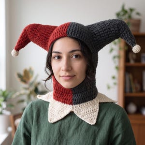 Puede incluir: Un gorro de bufón de ganchillo bicolor con un diseño dividido en rojo y gris oscuro, que presenta dos cuernos largos y puntiagudos rematados con pequeños pompones color crema y un cuello ancho y puntiagudo color crema.