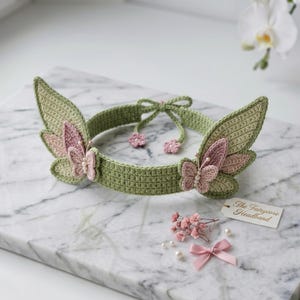 May include: A sage green crochet headband featuring two large wing-shaped side panels, each adorned with a small pink butterfly and white pearl accents. The band includes thin ties with flower-shaped ends and a tag reading The Fairycore Headband.