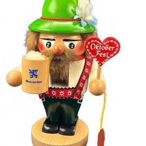 May include: A wooden nutcracker figure wearing a green hat with a blue flower, holding a light wood beer mug with a blue lion logo and the word Bavarian, and a red heart-shaped sign with the text Oktoberfest, standing on a round base.