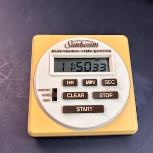 May include: A square Sunbeam Electronic Time Keeper with a yellow outer casing and a white circular face. The digital display shows 11:50:33 AM. It features buttons labeled HR, MIN, SEC, CLEAR, STOP, and START, plus a sliding switch for settings.