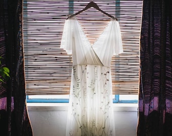 May include: A white maxi dress with floral embroidery hanging in a window. The dress has a V-neckline and short sleeves.