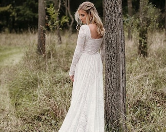 May include: A long, white lace wedding dress with long sleeves and a square neckline. The dress has a flowing skirt that trails on the ground. The person is standing in a forest setting with tall trees and tall grass.