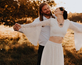 Puede incluir: Una pareja posa para una foto de boda en un campo. La mujer lleva un vestido blanco de encaje con un top corto y mangas largas. El hombre lleva una camisa blanca y pantalones negros. La pareja sonríe y se mira.