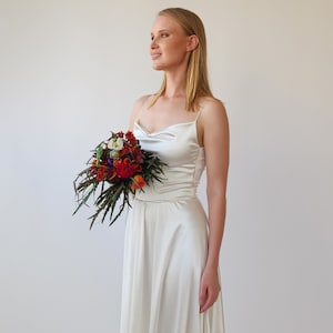 May include: A white satin wedding dress with a cowl neck and spaghetti straps. The dress has a flowing skirt and is paired with a bouquet of colorful flowers.