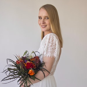 May include: A woman in a white lace wedding dress with a flowing skirt holds a bouquet of colorful flowers. The bouquet includes red, orange, and yellow flowers with green leaves.