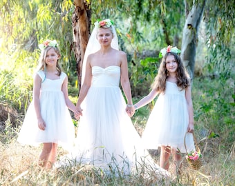May include: Three people wearing white dresses and flower crowns stand in a woodland setting. The person in the middle is wearing a veil. The two people on the sides are holding hands with the person in the middle.
