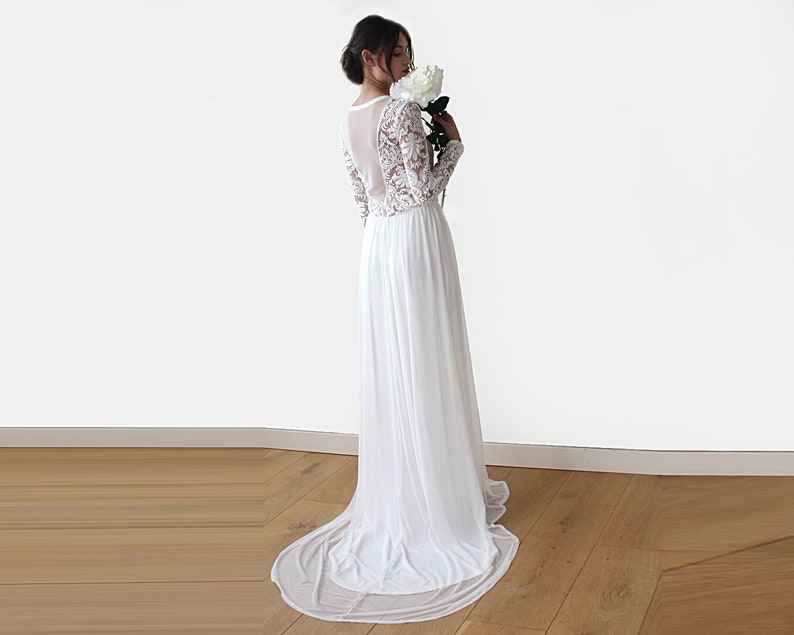Puede incluir: Un vestido de novia blanco de encaje de manga larga con una falda de gasa fluida. El vestido tiene un panel trasero transparente y un escote redondo.