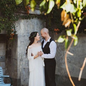 May include: A bride and groom stand close together, looking at each other. The bride is wearing a white lace dress and a floral crown. The groom is wearing a white shirt, a black vest, and black pants. They are standing in front of a gray wall with green vines growing on it.