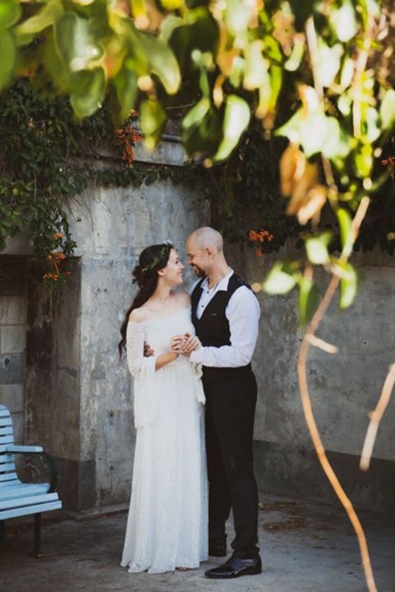 May include: A bride and groom stand close together, looking at each other. The bride is wearing a white lace dress and a floral crown. The groom is wearing a white shirt, a black vest, and black pants. They are standing in front of a gray wall with green vines growing on it.