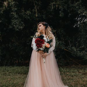 Puede incluir: Una novia con un vestido de novia de tul color melocot&oacute;n sostiene un ramo de flores rojas, blancas y melocot&oacute;n. Est&aacute; de pie frente a un fondo verde oscuro frondoso.