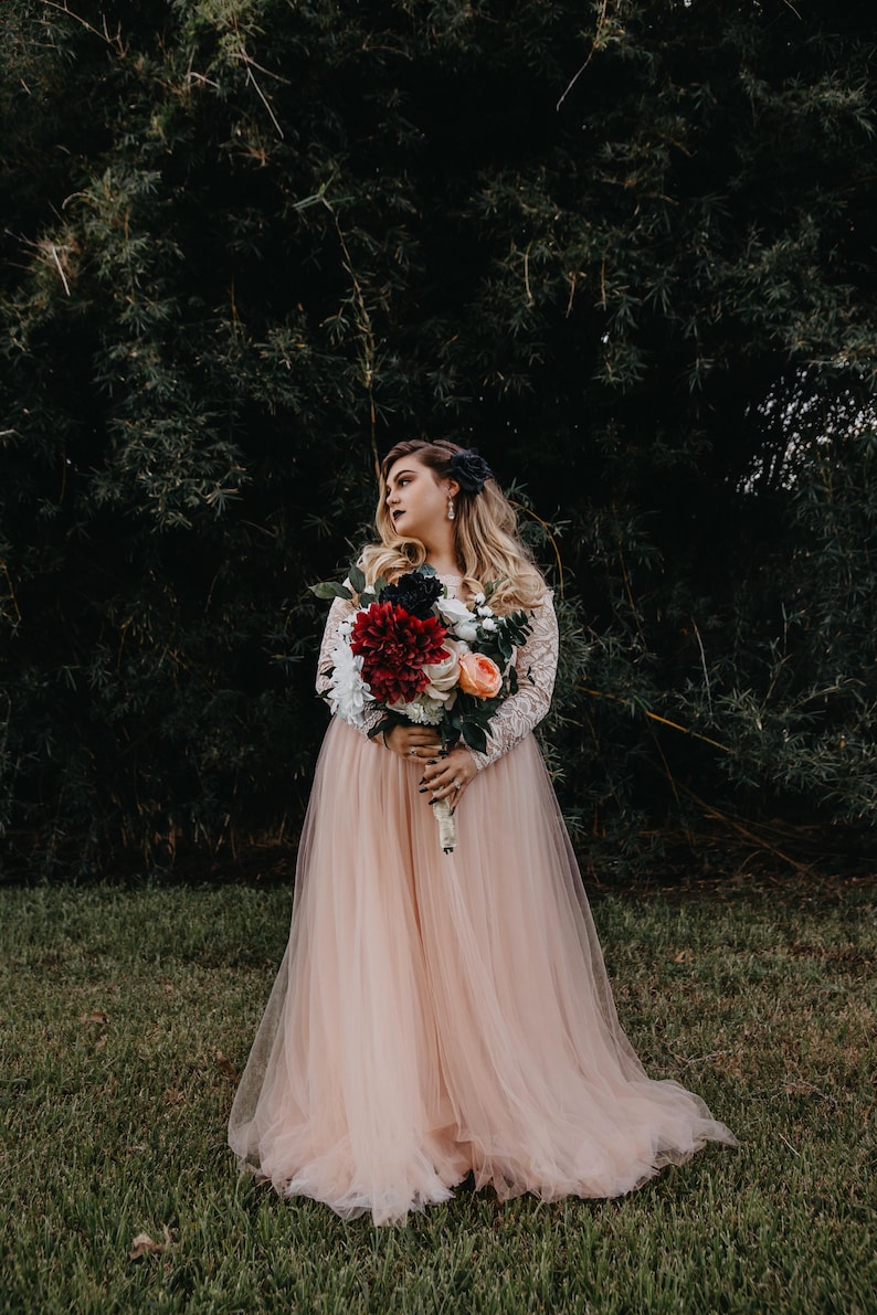 Puede incluir: Una novia con un vestido de novia de tul color melocot&oacute;n sostiene un ramo de flores rojas, blancas y melocot&oacute;n. Est&aacute; de pie frente a un fondo verde oscuro frondoso.