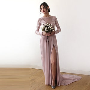 Puede incluir: Un vestido maxi rosa polvoriento con mangas largas y cuello alto. El vestido tiene un corpi&ntilde;o de encaje transparente y una falda fluida con una abertura en el muslo. La modelo sostiene un ramo de flores blancas.