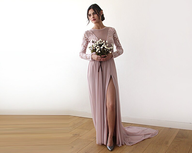 Puede incluir: Un vestido maxi rosa polvoriento con mangas largas y cuello alto. El vestido tiene un corpi&ntilde;o de encaje transparente y una falda fluida con una abertura en el muslo. La modelo sostiene un ramo de flores blancas.