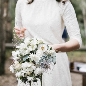 May include: A bride in a white lace dress holds a bouquet of white flowers with green foliage. The bouquet is made up of white flowers with green leaves and stems.