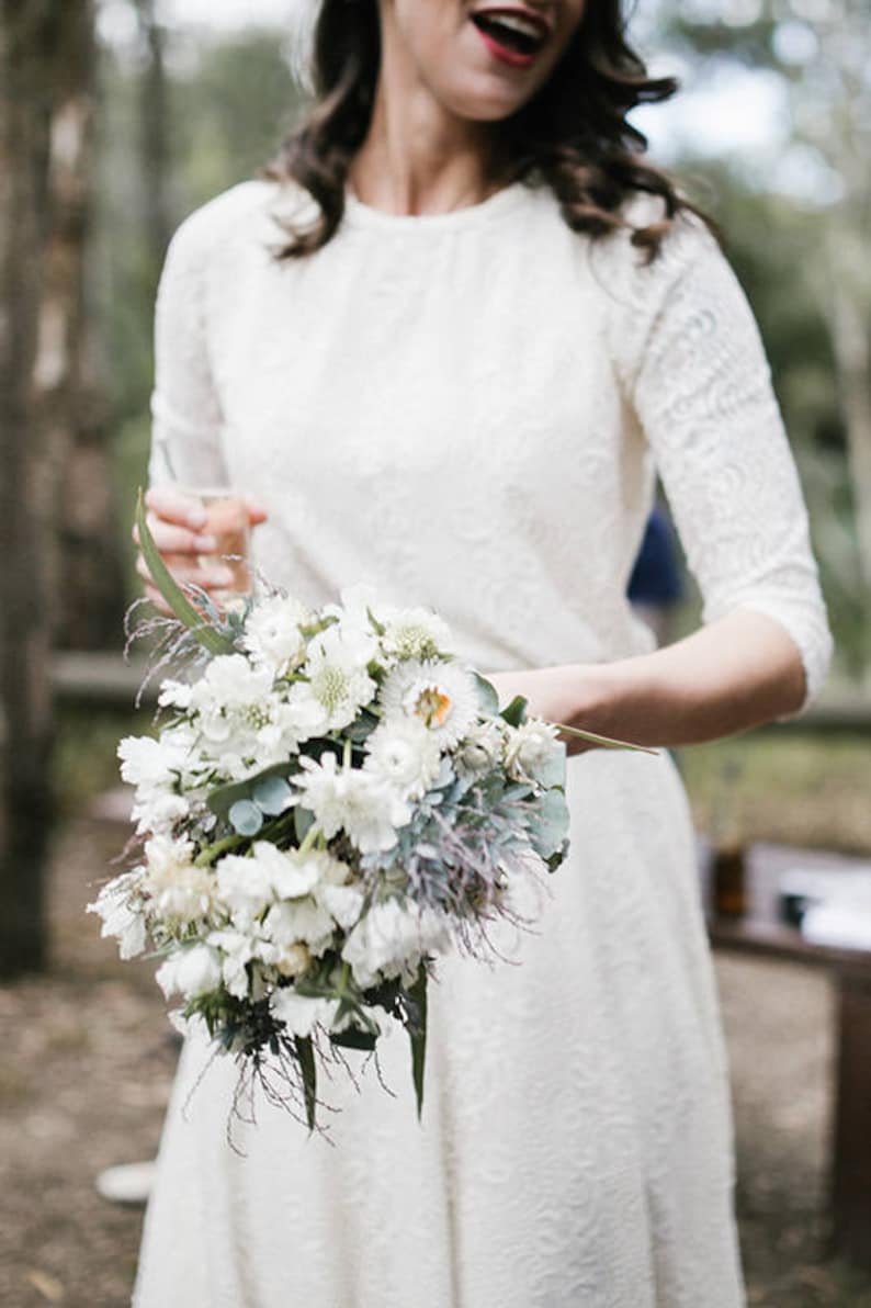 May include: A bride in a white lace dress holds a bouquet of white flowers with green foliage. The bouquet is made up of white flowers with green leaves and stems.