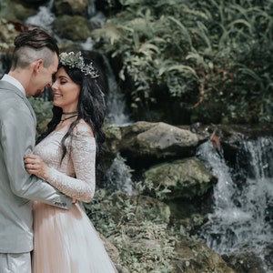 Puede incluir: Una pareja con atuendo de boda se encuentra de pie cerca uno del otro frente a una cascada. La mujer lleva un vestido blanco de encaje con una corona de flores y el hombre lleva un traje gris.