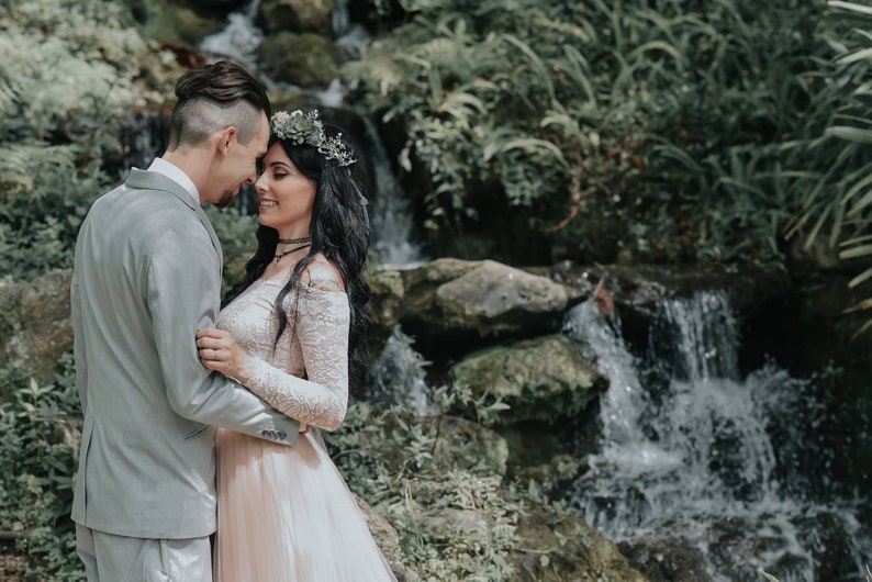 Puede incluir: Una pareja con atuendo de boda se encuentra de pie cerca uno del otro frente a una cascada. La mujer lleva un vestido blanco de encaje con una corona de flores y el hombre lleva un traje gris.