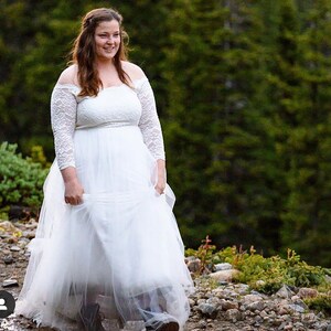 Puede incluir: Una novia con un vestido de novia blanco de encaje de hombros descubiertos con una falda de tul y mangas de encaje. Est&aacute; caminando por un sendero rocoso con vegetaci&oacute;n verde en el fondo.