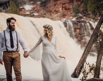 Op de afbeelding: Een stel houdt elkaar vast voor een waterval. De vrouw draagt een witte kanten jurk en een bloemenkrans. De man draagt een wit overhemd, een bruine broek en een bruine stropdas.